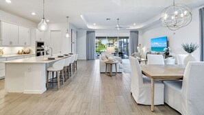 Una cucina moderna con isola centrale, zona pranzo con tavolo in legno e sedie bianche, e un soggiorno con divano e televisore.