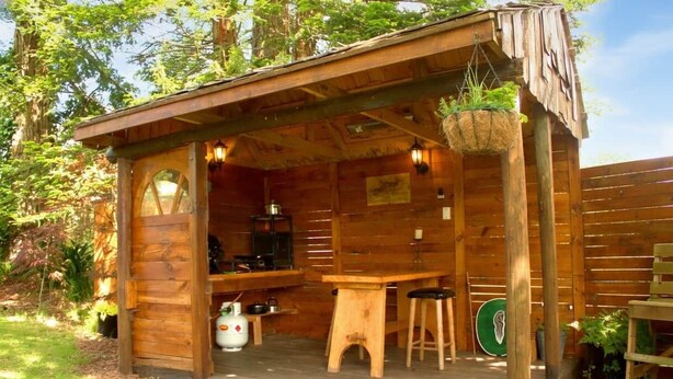A rustic wooden cabin with a porch, a small kitchen area, and a hanging plant. - A romantic night in a Hole in the Ground (Tauwhare)