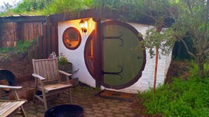 A small, rustic wooden structure with a green door and a circular window, surrounded by greenery and a wooden bench. - A romantic night in a Hole in the Ground (Tauwhare)