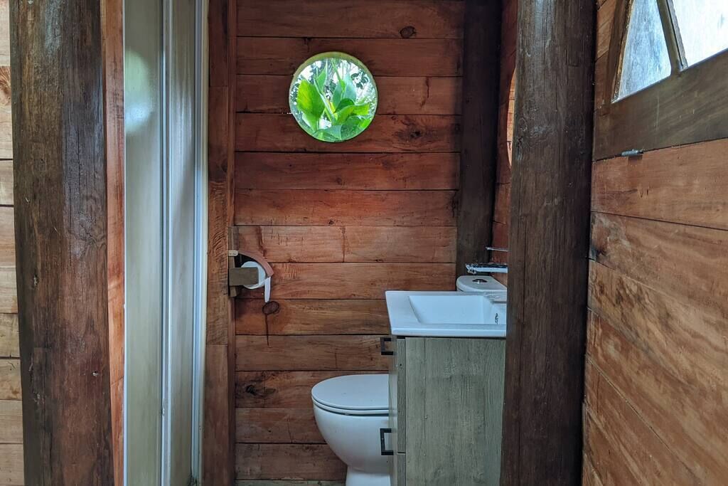 Ett badrum i en stuga, inrett med träpanel, och utrustat med toalett, handfat och ett fönster med utsikt över grönskan.