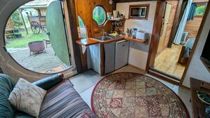 A cozy, rustic kitchen with a wooden interior, a sink, and a microwave. A large round window offers a view of the outdoors. - A romantic night in a Hole in the Ground (Tauwhare)