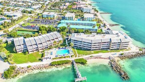Vista aérea de un complejo turístico costero con varias edificaciones, una piscina y un muelle que se adentra en el agua.