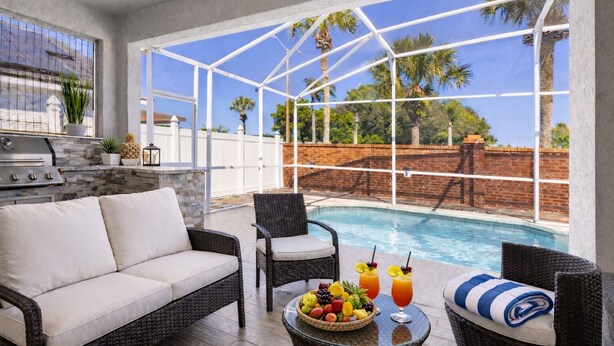 Un espace aménagé au bord de la piscine, avec un canapé, des fauteuils, une table garnie de fruits et de boissons, et une vue sur les palmiers.