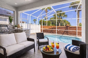 A poolside area with a sofa, chairs, a table with fruit and drinks, and a view of palm trees. - VG Luxury Home - Heated Pool close to Disney Parks (Davenport)