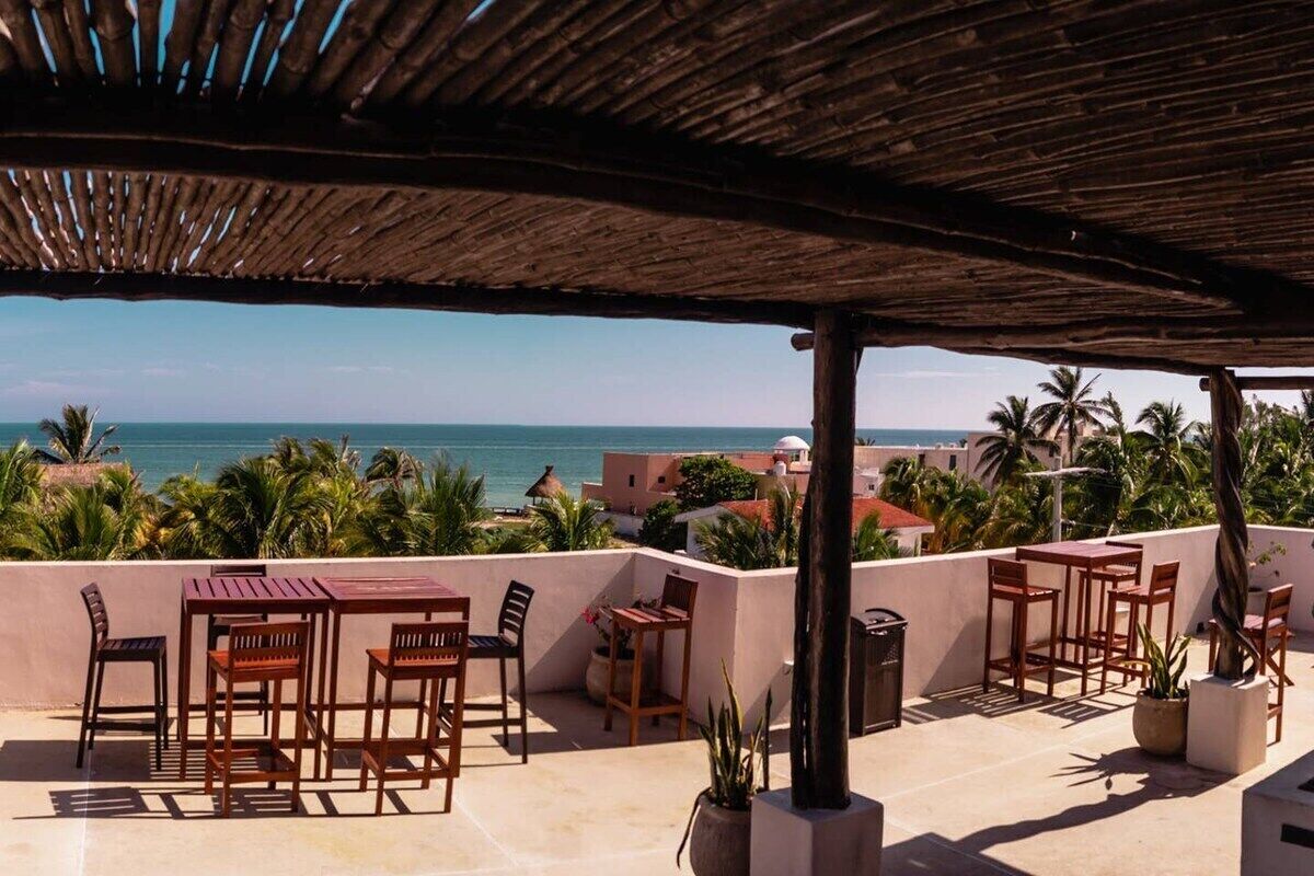A terrace with wooden furniture and potted plants, overlooking a coastal view with palm trees and buildings.