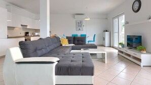 A modern living room with a grey sectional sofa, a white armchair, a coffee table, and a TV on a stand. There is a kitchen area in the background with white cabinets and a microwave.