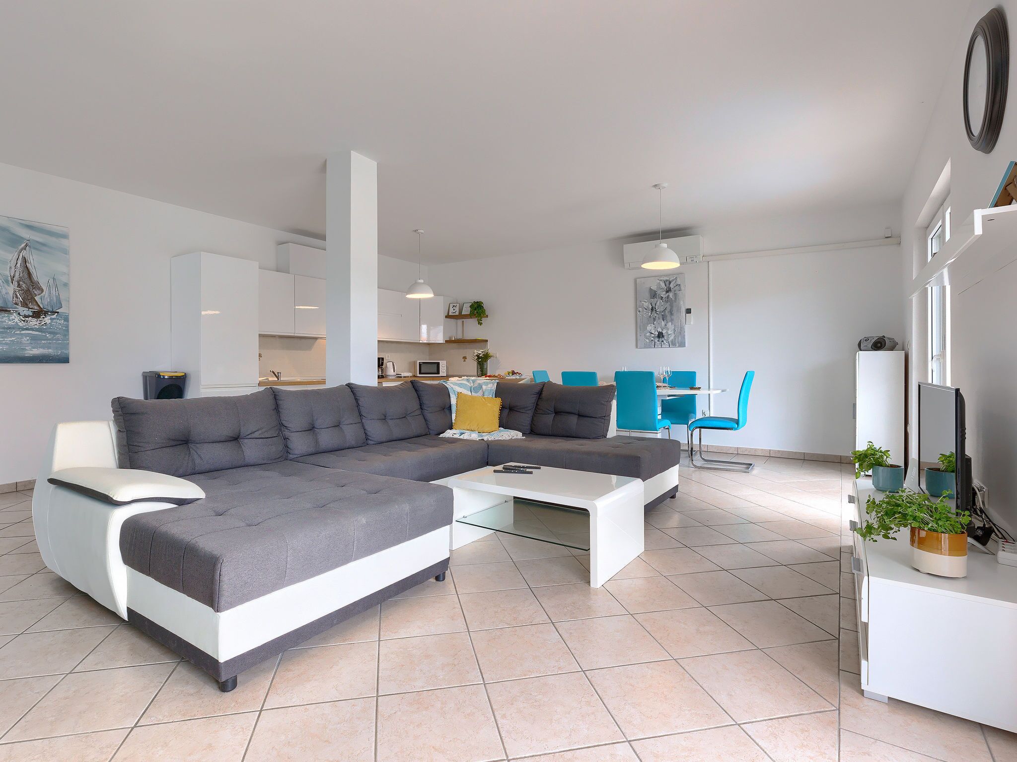 Un salon moderne avec un canapé d’angle gris, une table basse blanche et des chaises bleues. On aperçoit une cuisine en arrière-plan, avec des meubles blancs, et un coin repas avec une table blanche et des chaises bleues.