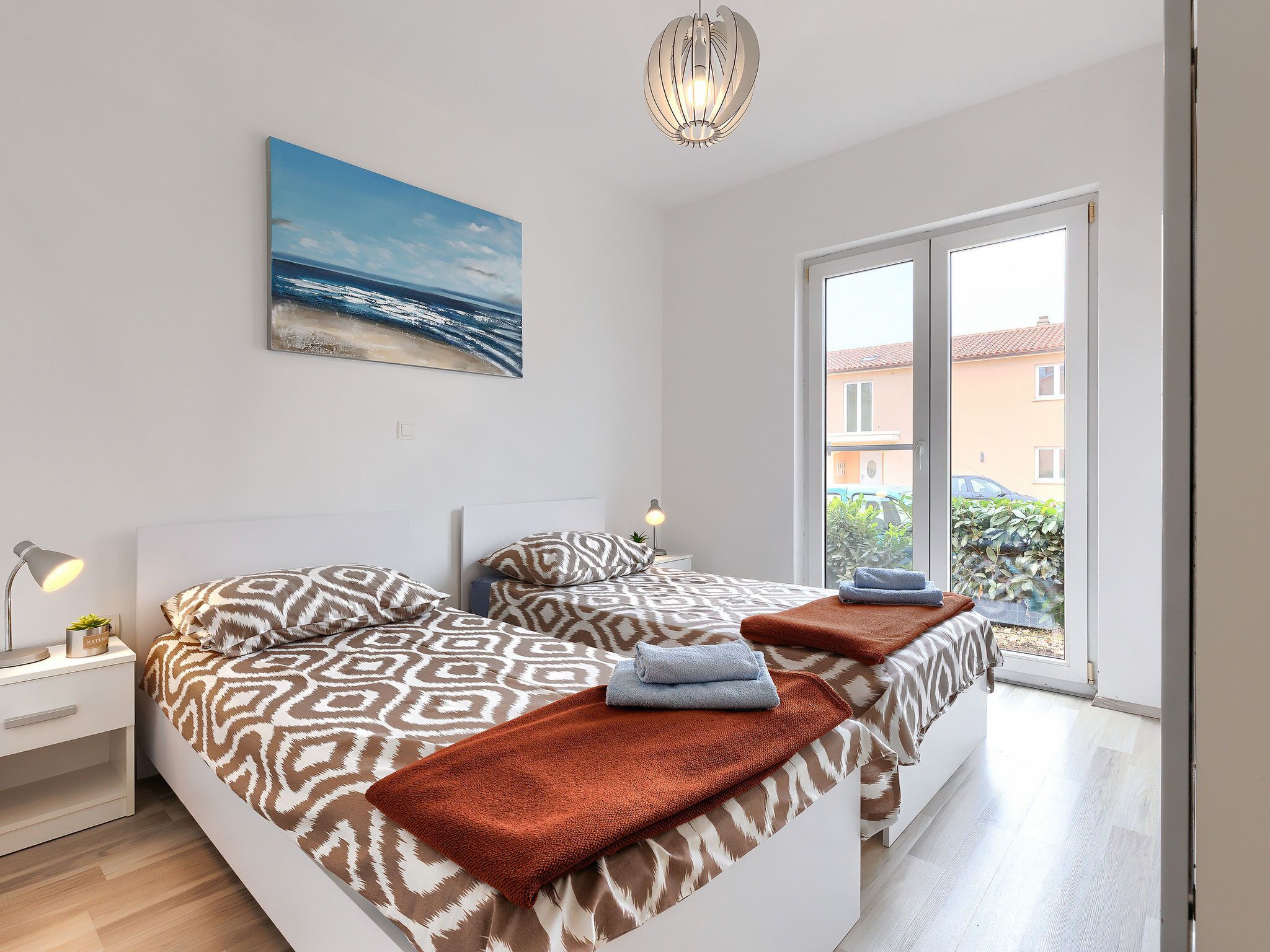 A modern bedroom with a large bed, a nightstand, a lamp, and a painting of a beach scene.
