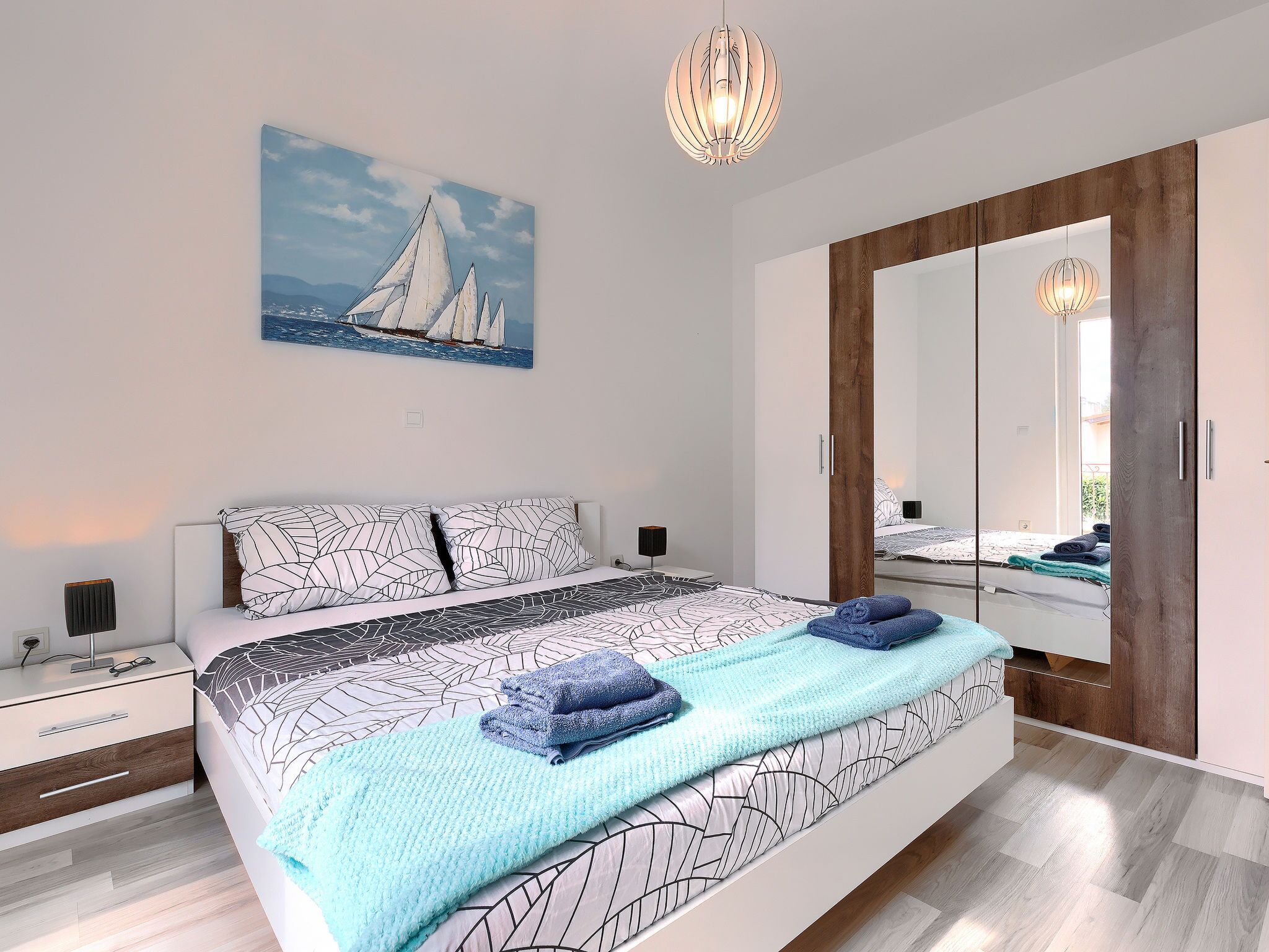 A modern bedroom with a bed, bedside tables, a large mirror, and a painting of sailboats.