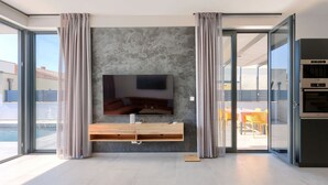 A modern living room with a flat-screen TV mounted on a textured wall, a wooden console table, and a sliding glass door leading to an outdoor patio. - Villa MI by Interhome (Pula)