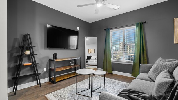 A modern living room with a grey sofa, a flat-screen TV, a ceiling fan, and a window with green curtains. - The Platinum Suite Penthouse The Gulch  (Nashville)