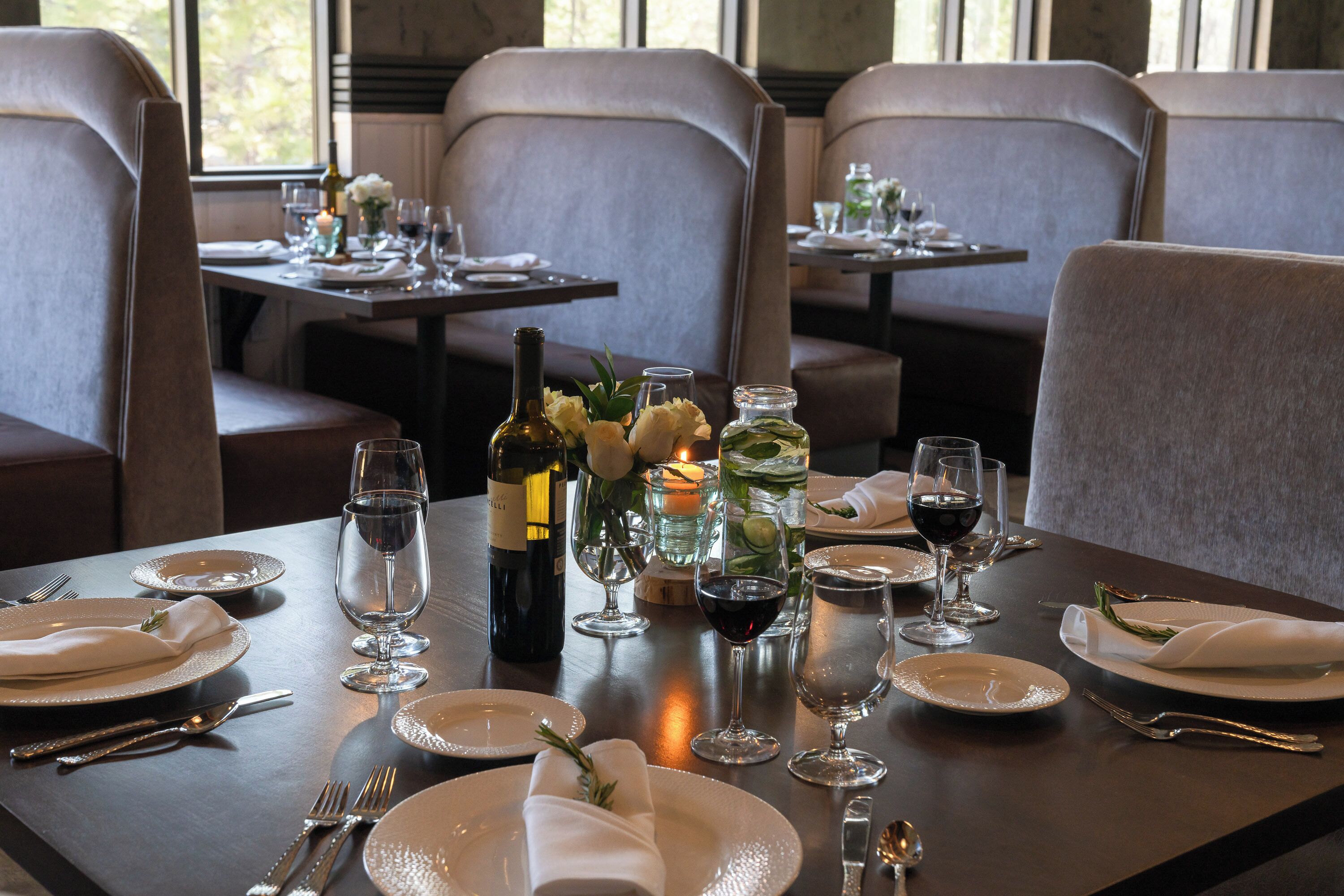 A dining table set with plates, cutlery, glasses, and a bottle of wine, with a candle and flowers on the table.