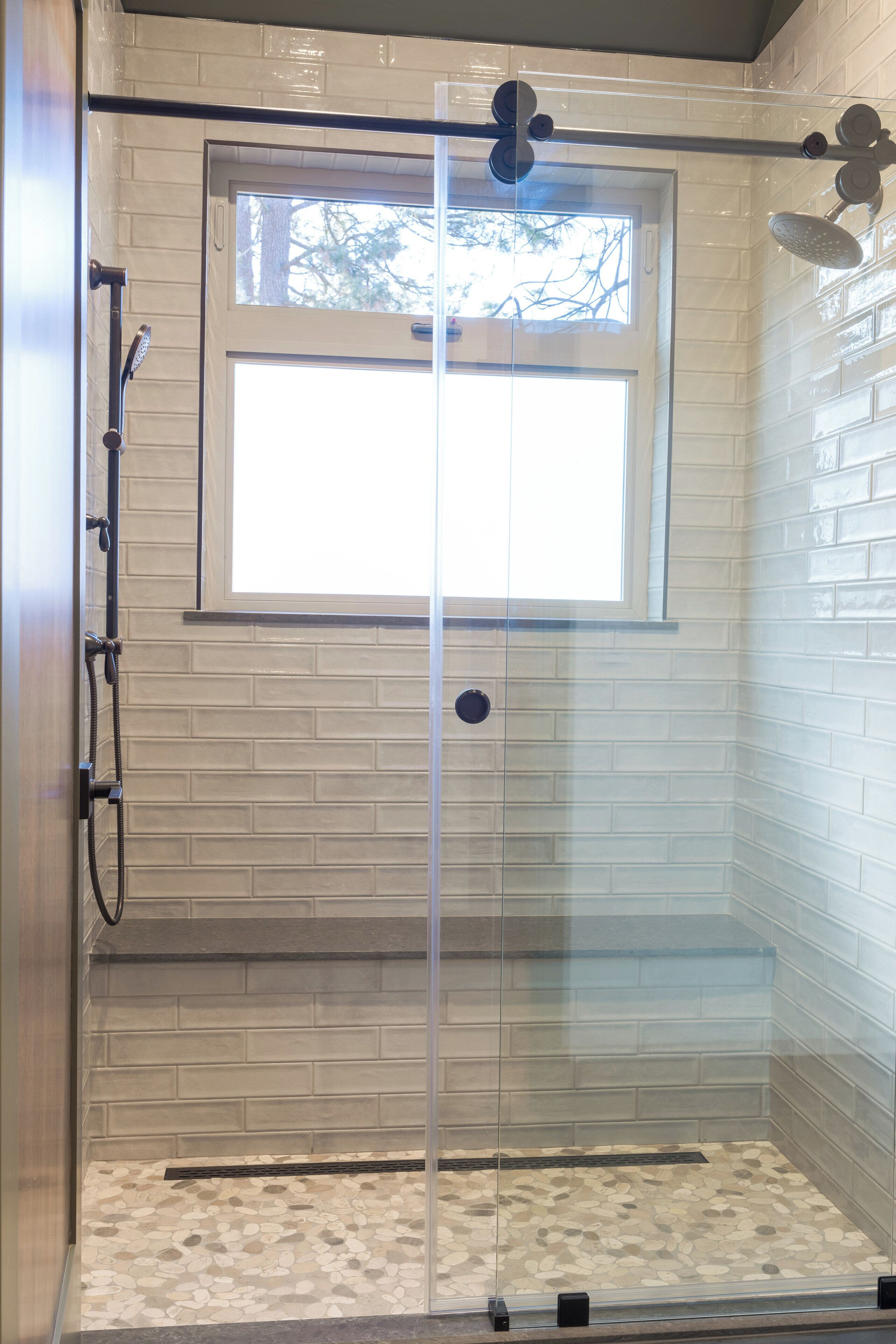 A glass-enclosed shower with a window, white brick walls, and a pebble-patterned floor.