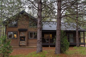 A rustic cabin with a stone fireplace, large windows, and a deck surrounded by trees.