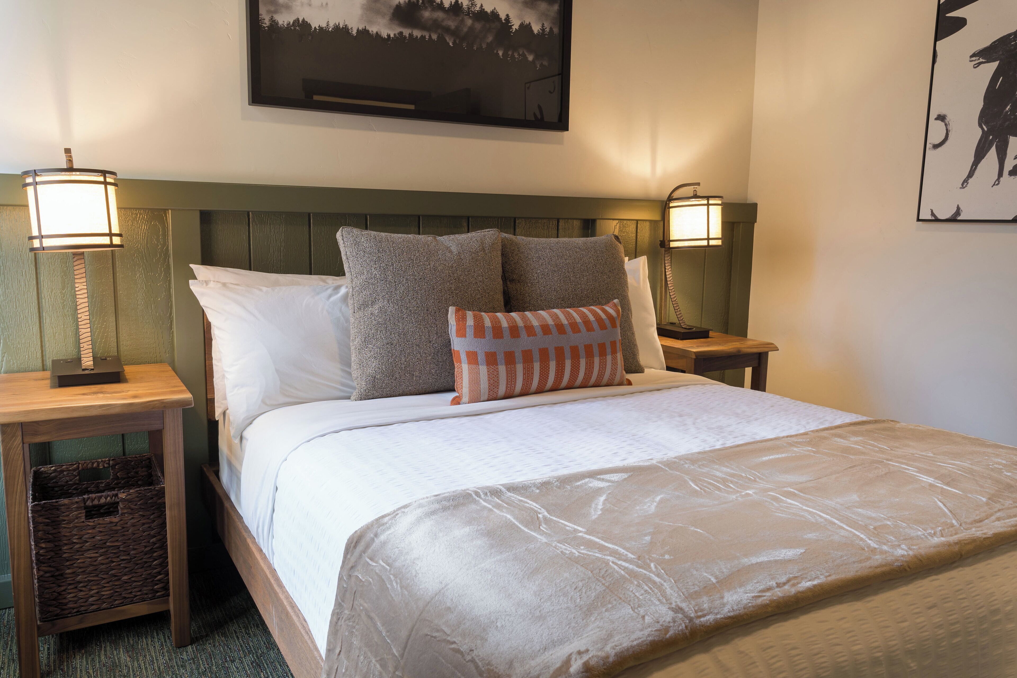A neatly made bed with a white comforter and two pillows, flanked by two bedside tables with lamps, a framed picture above the bed, and a wooden headboard.