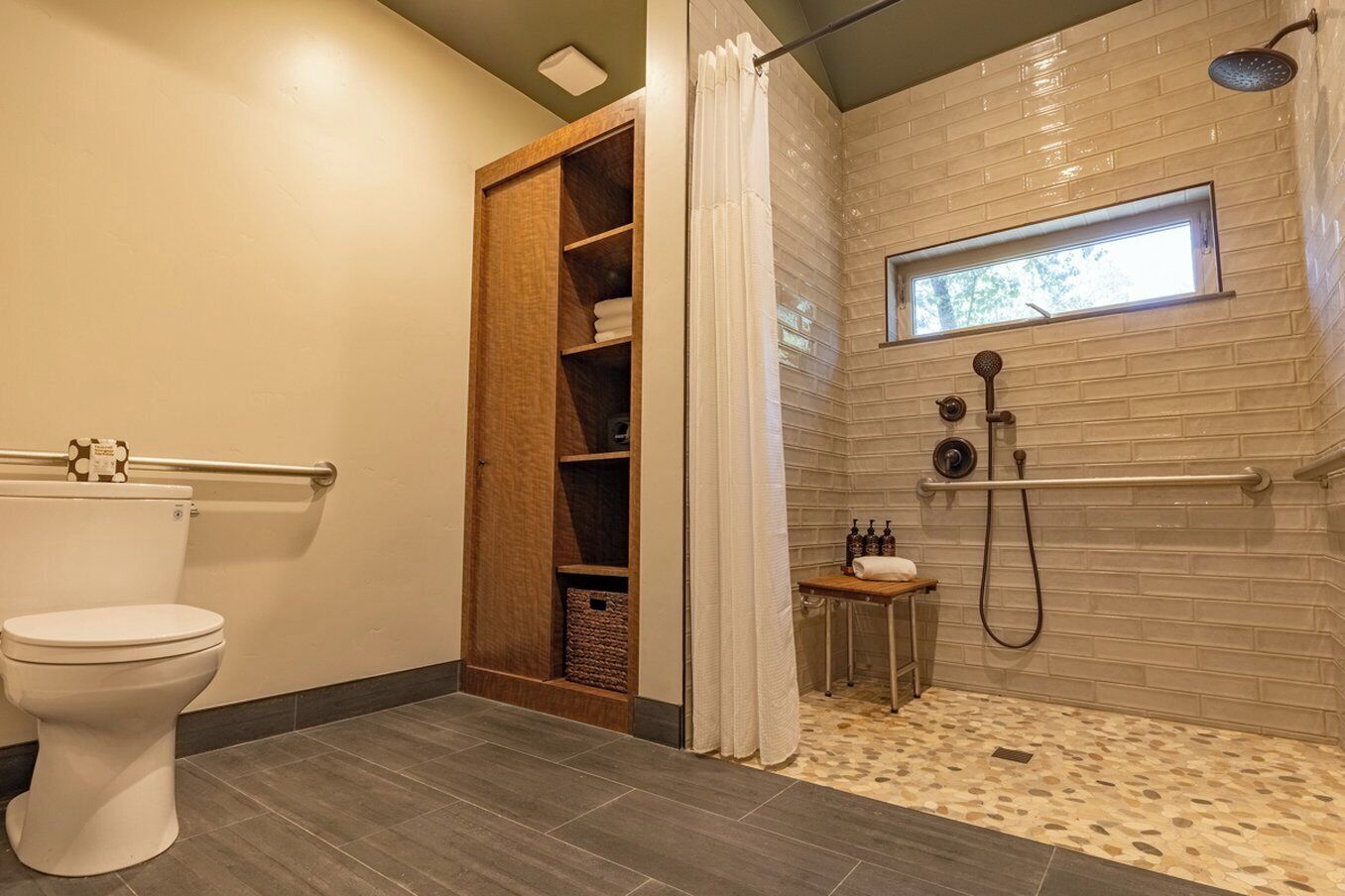 A bathroom with a toilet, a wooden cabinet, a shower area with a window, and a small sink.