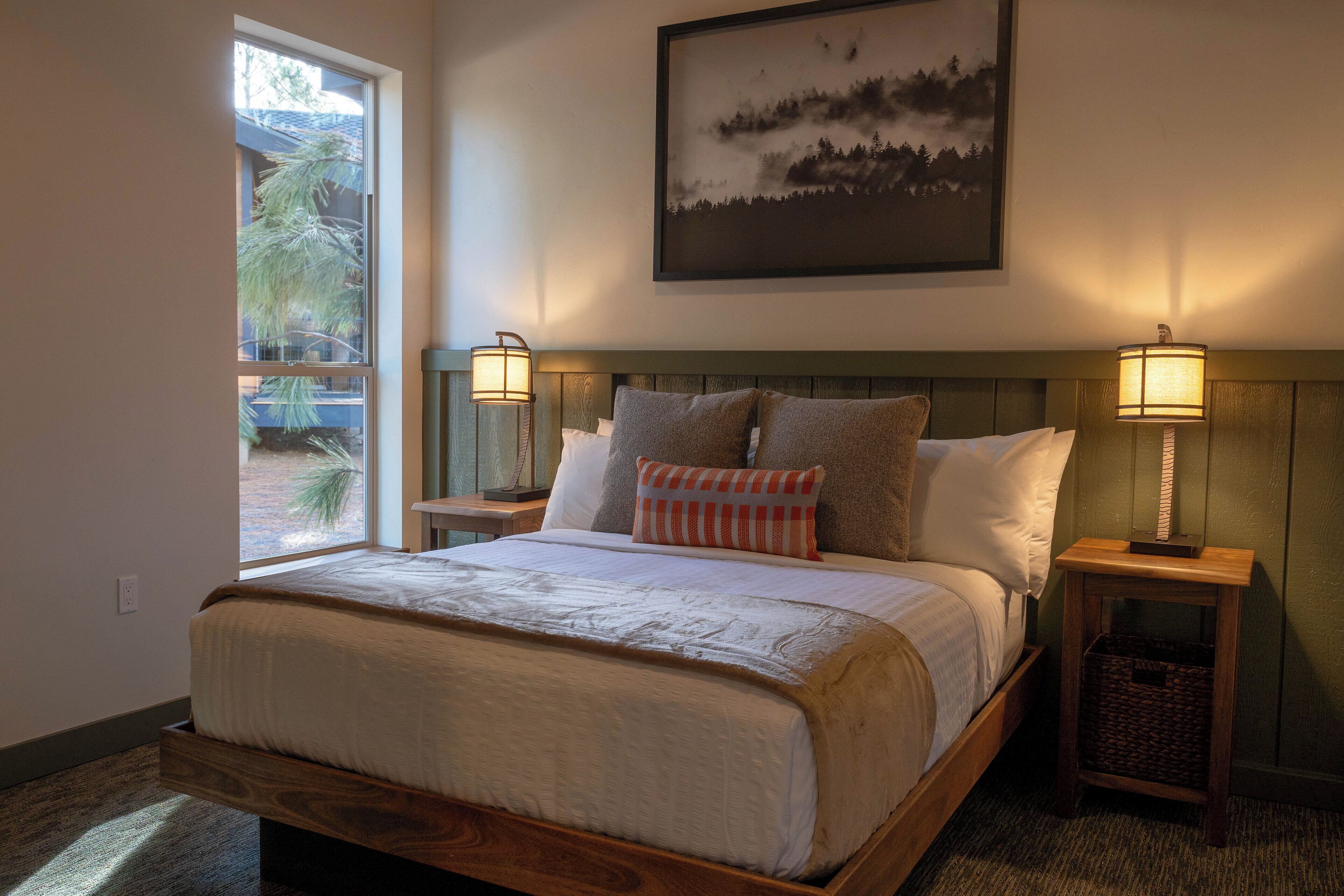 A bedroom with a wooden bed, two bedside tables with lamps, a window with a view, and a painting on the wall.