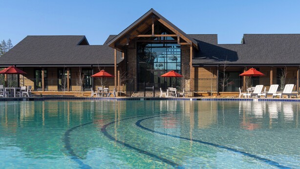 A clear swimming pool with lounge chairs and umbrellas, a building with a large glass window, and a wooden exterior.