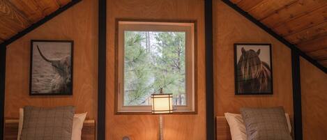 A wooden cabin room with two beds, a window with a view of trees, and framed artwork on the walls.