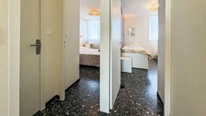 A corridor leading to a bedroom with a bed, a bedside table, and a lamp. The floor is tiled, and there is a door on the left. - Le Mireille by Interhome (Le Grau du Roi)