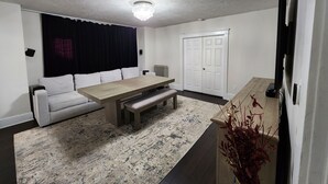 A modern living room with a sofa, a wooden dining table, a bench, a radiator, a white door, and a television. - Spacious and luxurious 3-bedroom home w/jetted bathtub (Worcester)