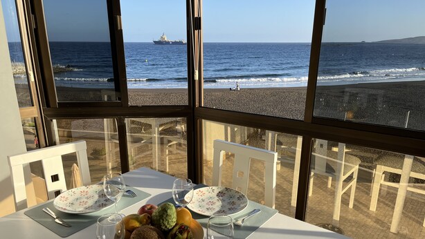 A table set for two with a fruit bowl, wine glasses, and plates, overlooking a beach with a ship in the distance.