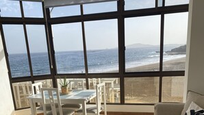 A room with a large window overlooking the beach, a white sofa, a glass coffee table, and a small table with chairs.