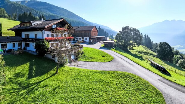 Ein traditionelles Chalet in den Alpen mit einer geschwungenen Auffahrt, umgeben von saftig grünen Wiesen und Bergen im Hintergrund.