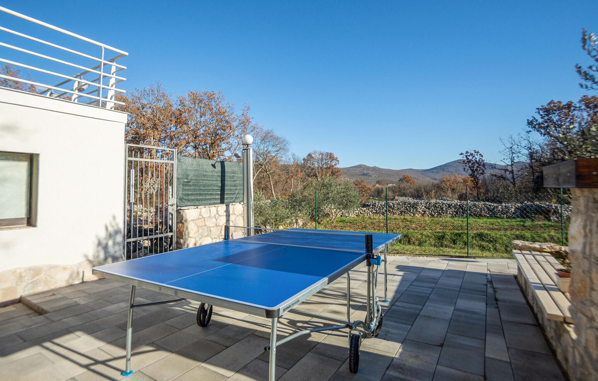 A ping pong table on a patio with stone flooring, a metal railing, and a view of trees and hills in the background.