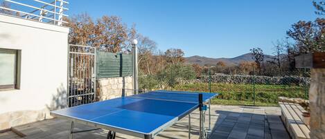 A ping pong table on a patio with stone flooring, a metal railing, and a view of trees and hills in the background.