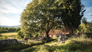 En bygning i sten med rødt tag, omgivet af frodigt grønt og en stenvæg.