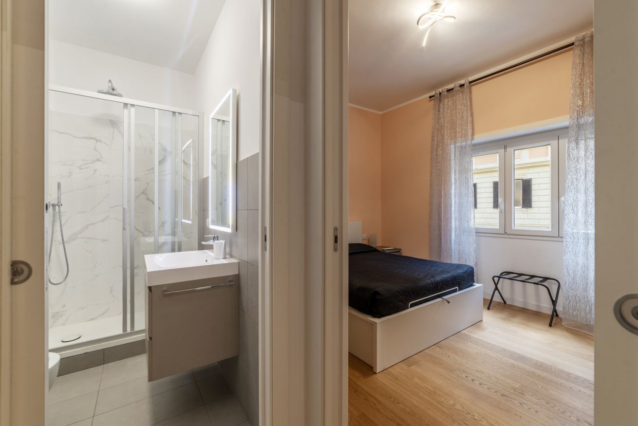 A bedroom with a bed, a small bedside table, a window with curtains, and a bathroom with a shower and toilet.