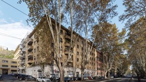 Un edificio de varios pisos con balcones, coches aparcados y árboles bordeando la calle.