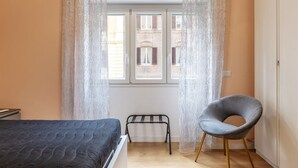 Un dormitorio con cama, una silla, un banco y una ventana con cortinas.