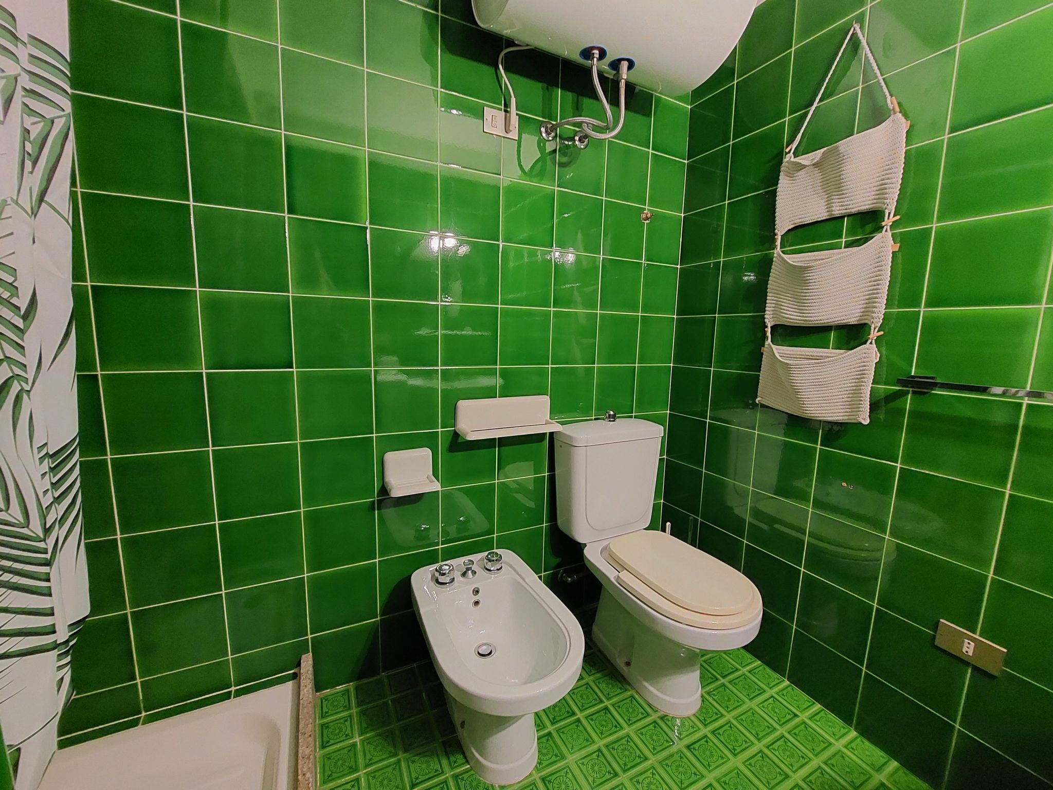 Une salle de bain avec des carreaux verts, des toilettes, un bidet et un panier suspendu.