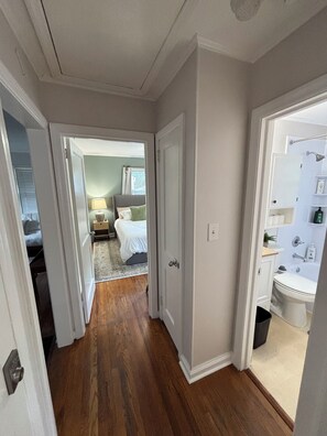 Un couloir avec un plancher en bois, une chambre avec un lit et une table de chevet, et une salle de bain avec un lavabo et des toilettes.