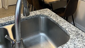 Une cuisine dotée d’un plan de travail en granit, d’un évier en acier inoxydable, d’une table à manger avec des chaises et d’une fenêtre avec des stores.