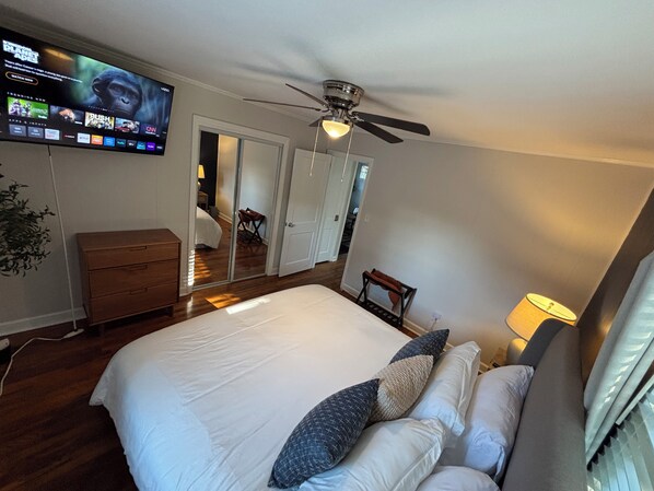A bedroom with a bed, a TV mounted on the wall, a ceiling fan, and a wooden dresser.