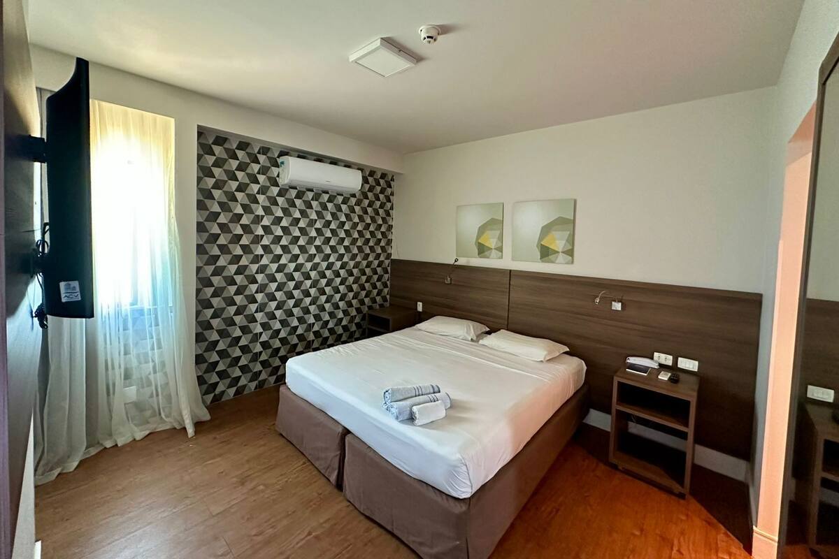A hotel room with a bed, a nightstand, an air conditioning unit, and a geometric-patterned wall.