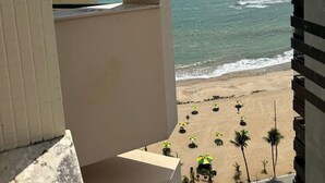Blick vom Balkon auf einen Strand mit Palmen und Sonnenschirmen, sowie ein Gebäude mit geometrischer Fassade.