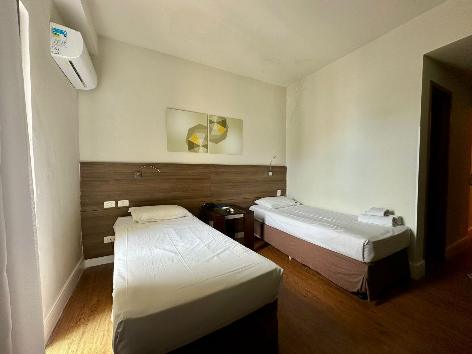 A hotel room with two single beds, a wooden headboard, a bedside table with a lamp, and a wall-mounted air conditioner.