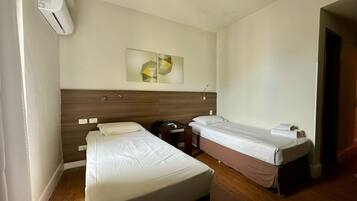 A hotel room with two single beds, a wooden headboard, a bedside table with a lamp, and a wall-mounted air conditioner.