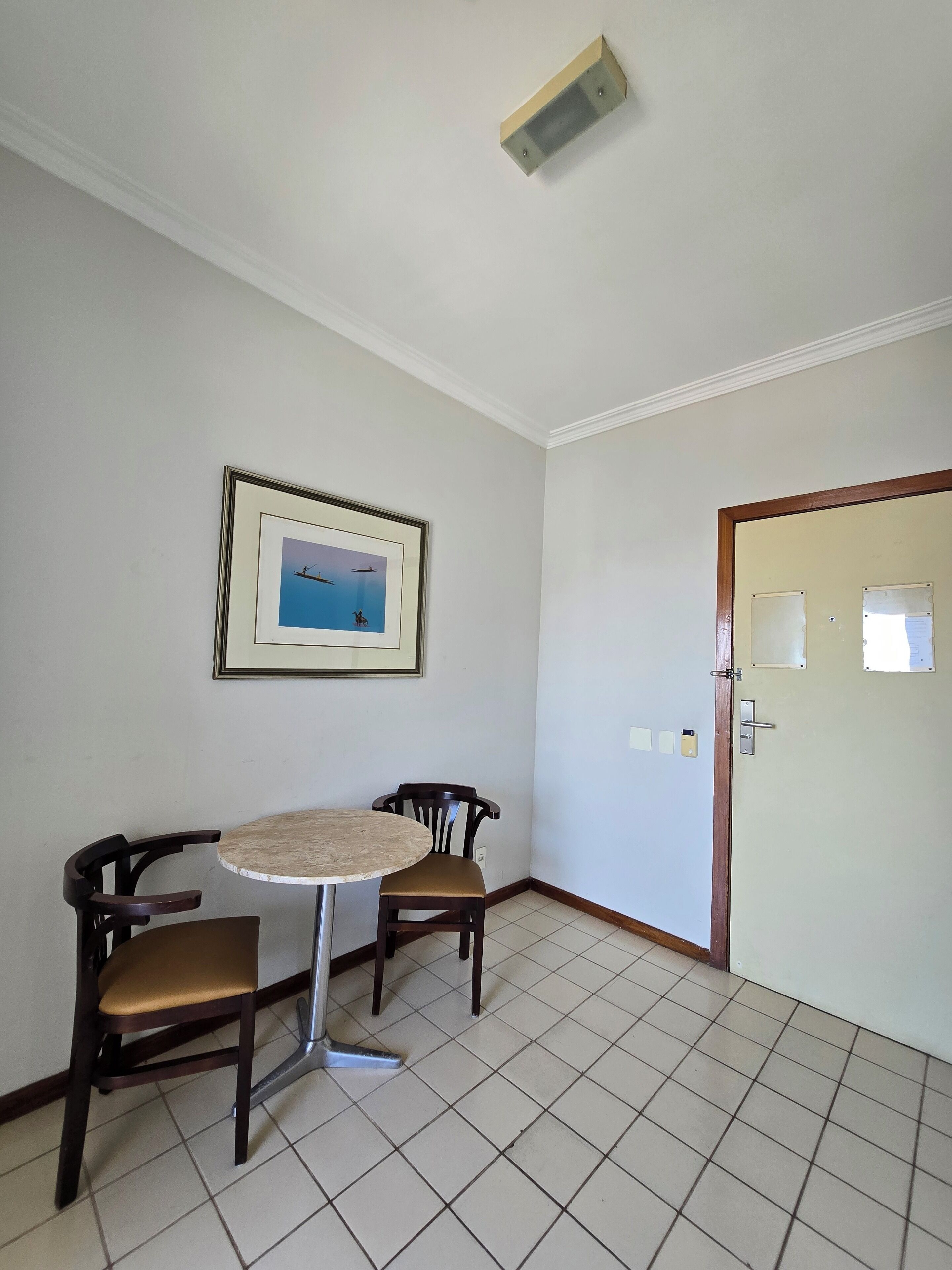 Una habitación con suelo de baldosas, una mesa redonda con dos sillas, un cuadro en la pared y una puerta con cerradura.