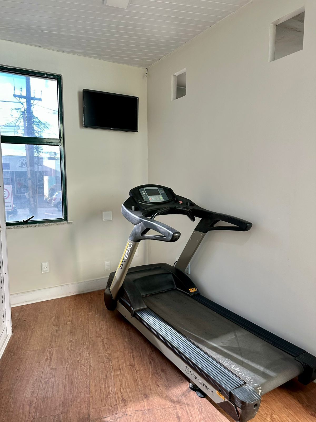Ein Laufband in einem Raum mit Fenster, einem an der Wand befestigten Fernseher und einem kleinen Fenster oberhalb des Laufbandes.