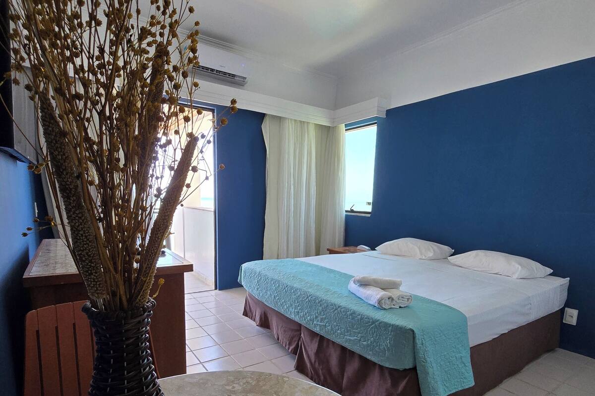 A hotel room with a bed, a table with a vase, and a window with curtains.