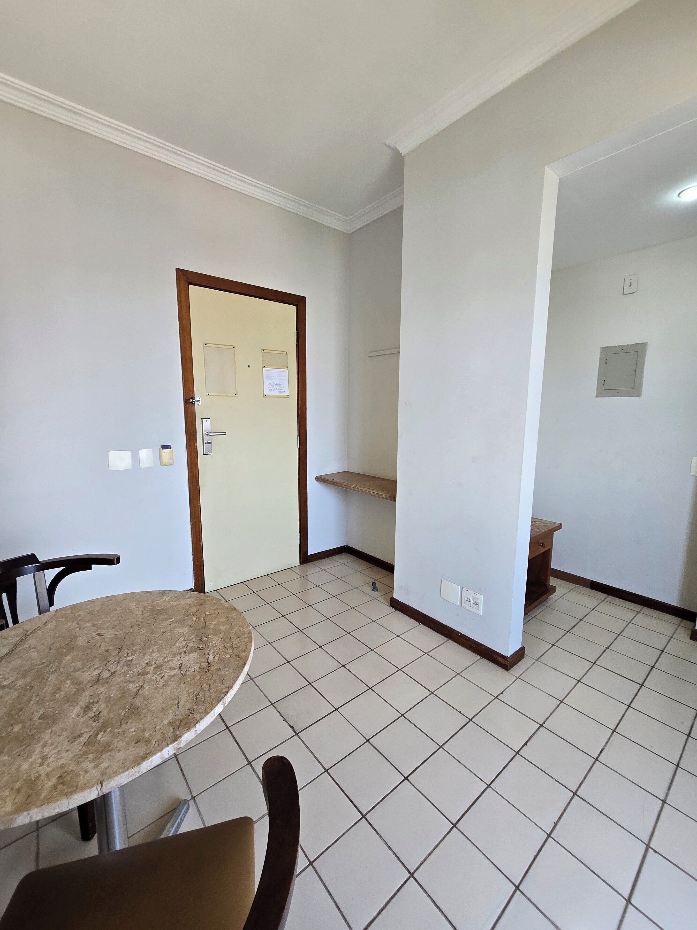 A room with a tiled floor, a round table with chairs, a door with a notice, and a shelf.