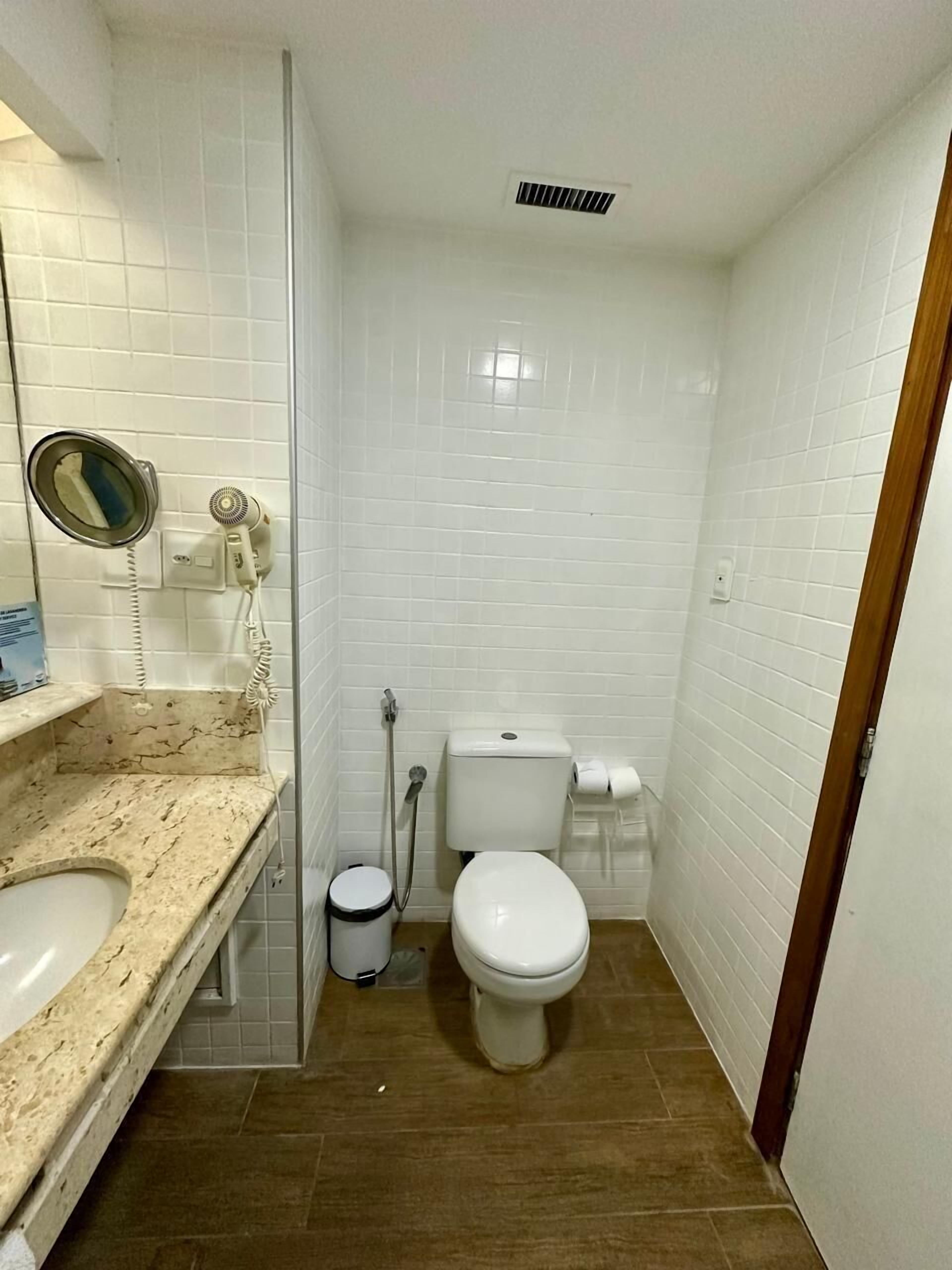 A small, clean bathroom with white tiles, a toilet, a sink, and a shower.