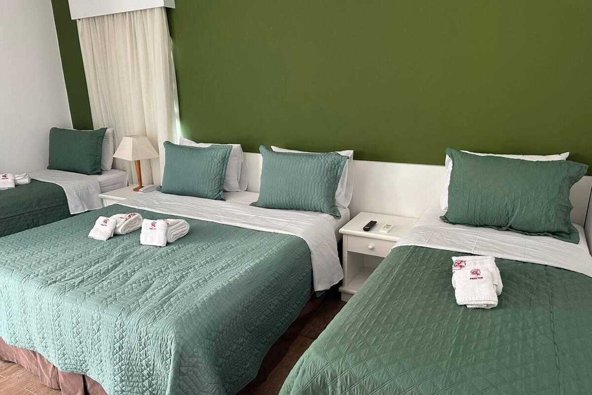 A hotel room with two beds, each with a green bedspread and matching pillows. There are white nightstands with lamps and a white dresser.