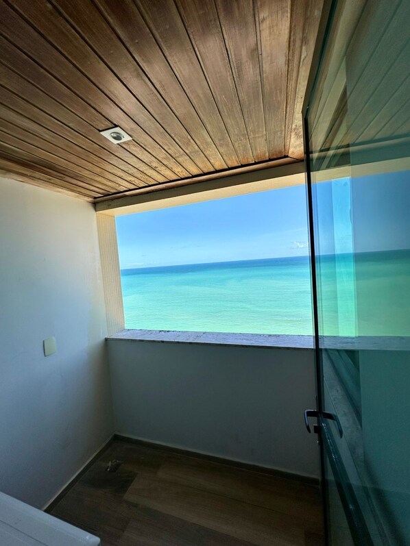 A room with a wooden ceiling, a window with a view of the ocean, and a glass door. -  Costa Mar Recife by ACM (Jaboatão dos Guararapes)