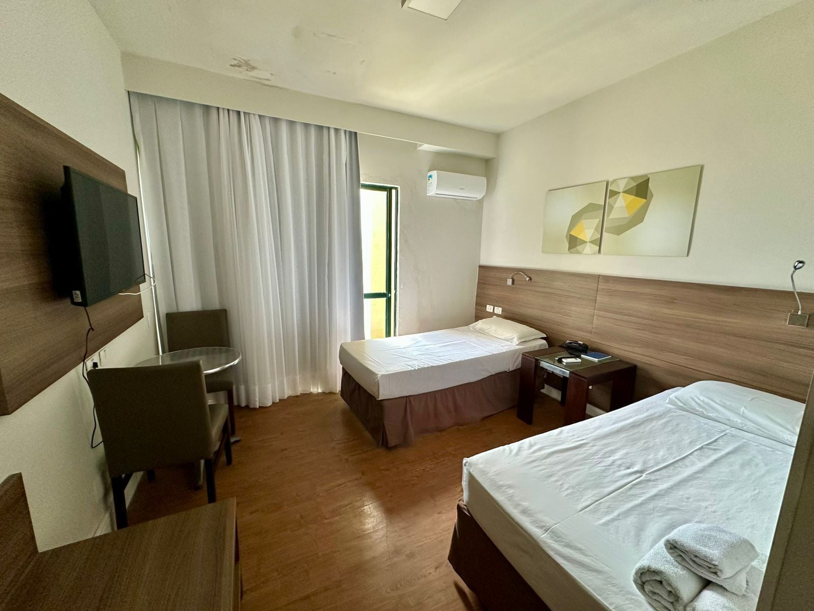 Hotelkamer met een bed, een bureau, een stoel, een televisie, een raam met gordijnen en een airconditioning.